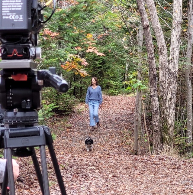 A camera films Karen as she walks in the forest with her dog